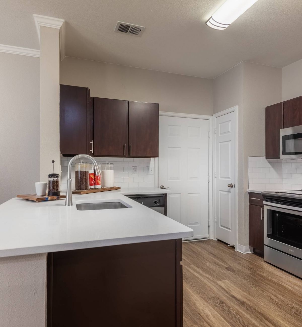 Brixton at Bear Creek modern kitchen with stainless steel appliances, dark wood cabinets, white countertops, and light wood flooring.