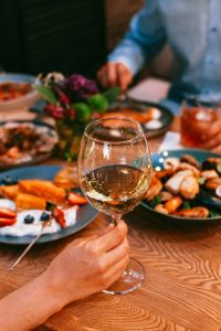Hand holding a glass of white wine over a wooden table with plates of food and blurred figures in the background.