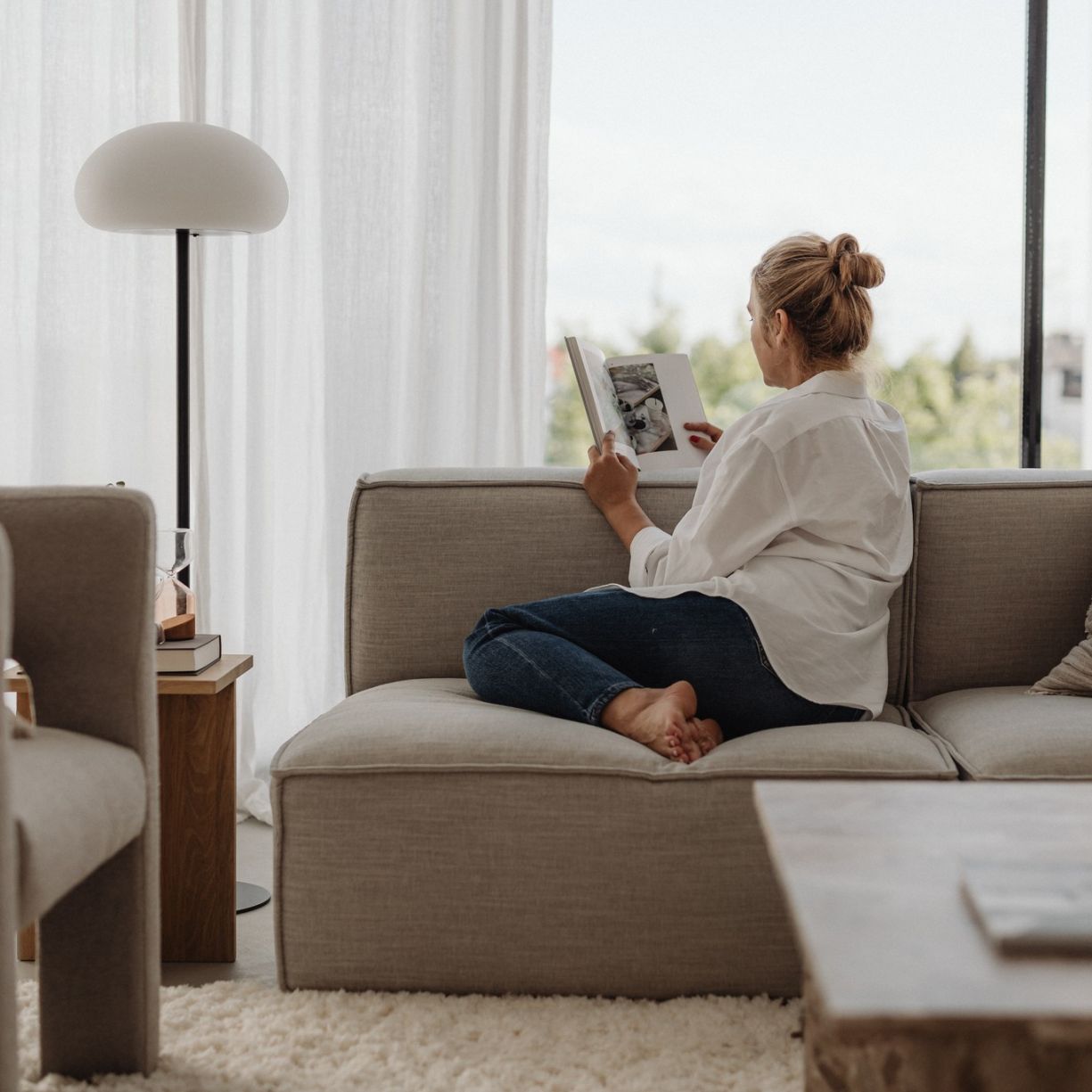 A woman sits on a sofa reading a book in a bright, modern living room with large windows and neutral-toned decor.