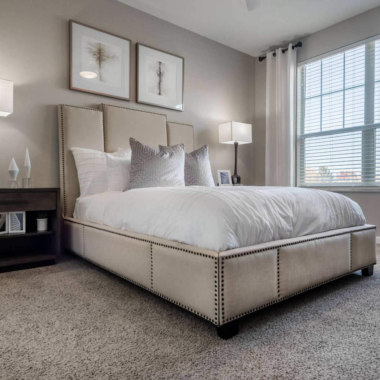 The Windsor elegant bedroom with a beige upholstered bed, bedside tables, two lamps, framed art, and a large window with curtains.