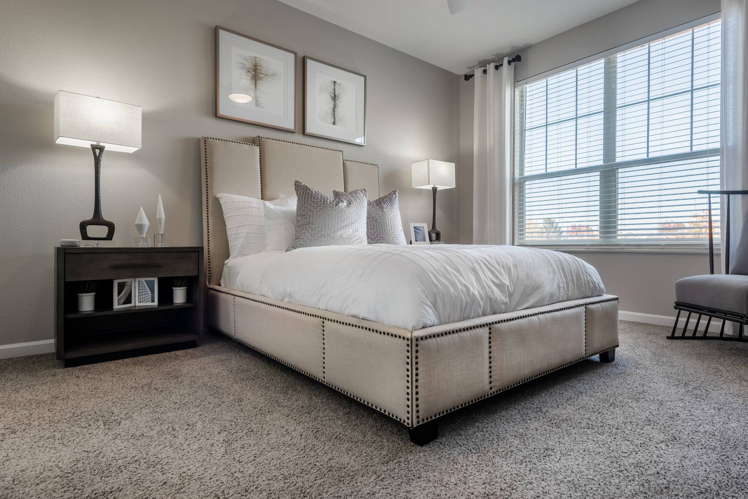 The Windsor elegant bedroom with a beige upholstered bed, bedside tables, two lamps, framed art, and a large window with curtains.
