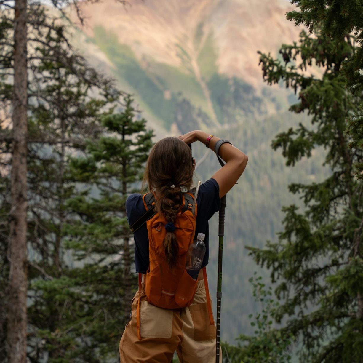 A person with a backpack stands among trees, taking a photo of a distant mountain under a cloudy sky.